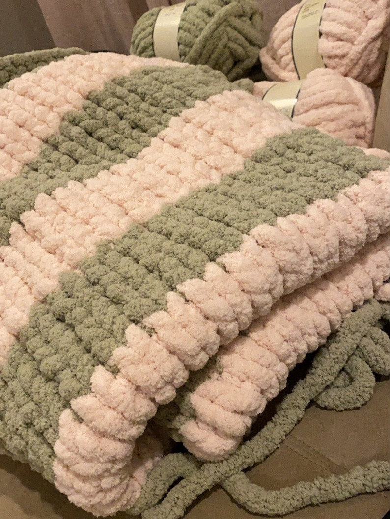 May include: A handmade, chunky knit blanket in a checkerboard pattern of soft pink and muted green. The blanket is made with thick, plush yarn, and several skeins of yarn are visible in the background.