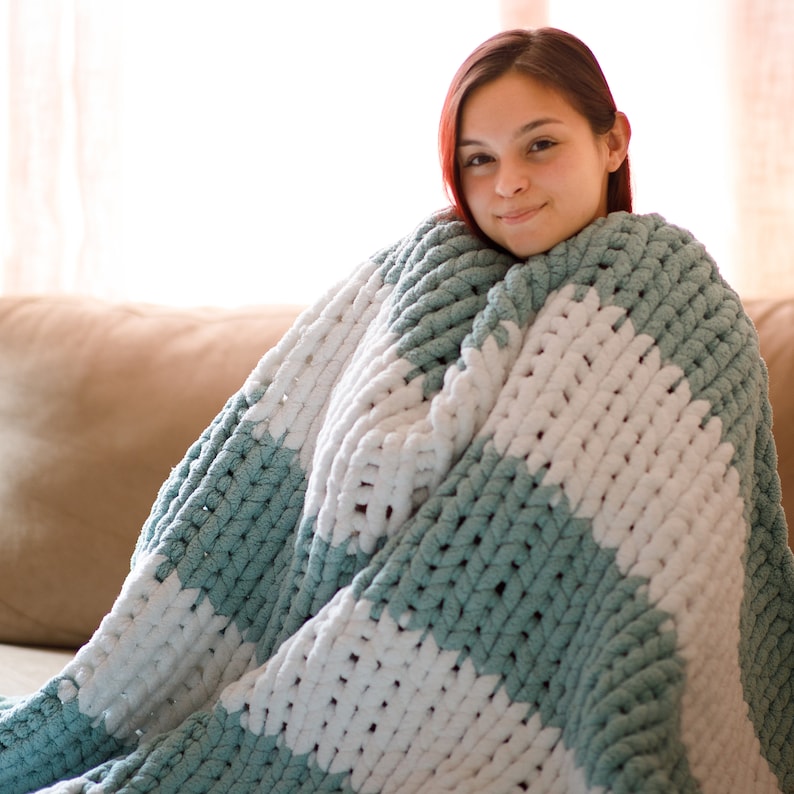 May include: A chunky knit blanket in a soothing color palette of white and a soft teal. The blanket features a striped pattern and is made of thick, plush yarn. This cozy throw is perfect for snuggling up on the couch or bed.