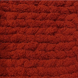 May include: Close-up of a textured, rust-colored fabric. The material has a looped, raised pattern, creating a plush, tactile surface. The color is a warm, earthy tone, suggesting comfort and a cozy feel. The fabric appears to be a rug or blanket.