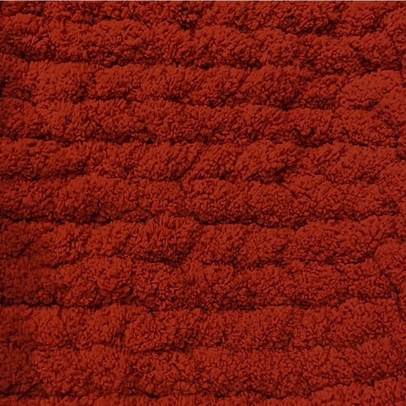 May include: Close-up of a textured, rust-colored fabric. The material has a looped, raised pattern, creating a plush, tactile surface. The color is a warm, earthy tone, suggesting comfort and a cozy feel. The fabric appears to be a rug or blanket.