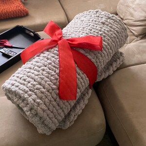 May include: A folded, light gray chunky knit blanket tied with a bright red satin ribbon. The blanket is made of thick, soft yarn and appears cozy. The ribbon is tied in a bow, suggesting it is a gift.