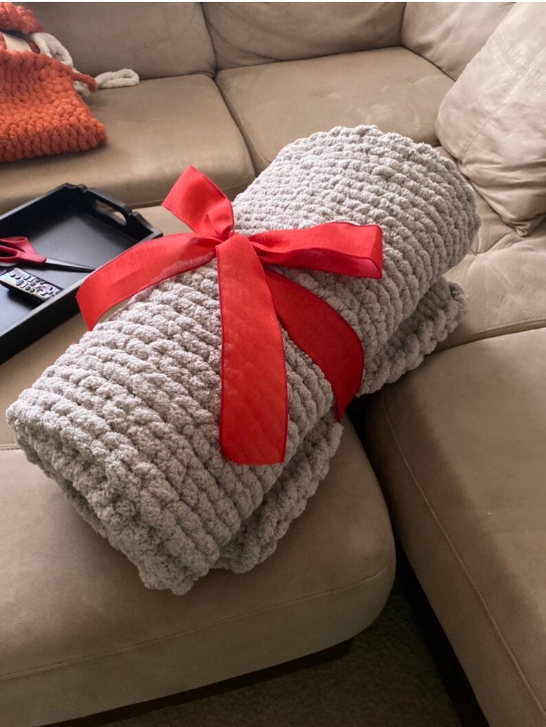 May include: A folded, light gray chunky knit blanket tied with a bright red satin ribbon. The blanket is made of thick, soft yarn and appears cozy. The ribbon is tied in a bow, suggesting it is a gift.