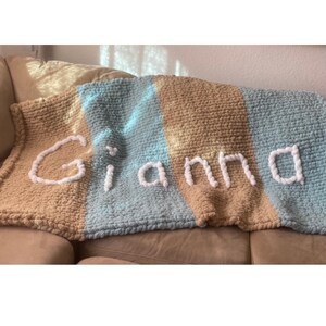 May include: A handmade, chunky knit blanket in tan, light blue, and blue stripes. The name "Gianna" is spelled out in white yarn across the blanket. The blanket is draped over a beige couch.
