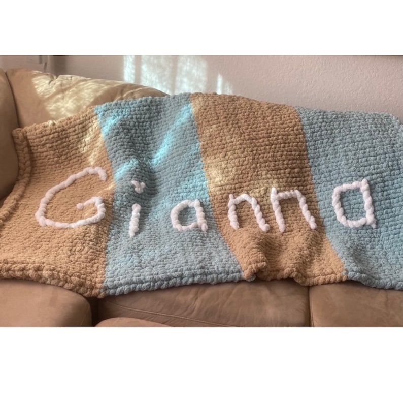 May include: A handmade, chunky knit blanket in tan, light blue, and blue stripes. The name "Gianna" is spelled out in white yarn across the blanket. The blanket is draped over a beige couch.