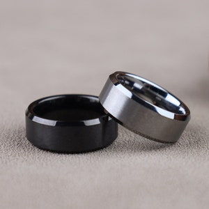 May include: Two tungsten carbide rings, one black and one silver, on a gray textured surface.