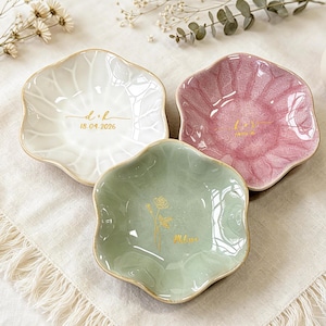 K&ouml;nnte beinhalten: Drei blumenf&ouml;rmige Schmuckschalen aus Keramik in Wei&szlig;, Rosa und Salbeigr&uuml;n mit einer rissigen Glasuroberfl&auml;che. Jede Schale ist mit goldfarbenen Schriftz&uuml;gen und Motiven verziert, darunter "d+k" mit "18.04.2026" sowie der Name "Melanie".