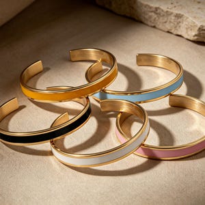 May include: A collection of five gold-toned metal cuff bracelets, each featuring a smooth, coloured enamel inlay. The bracelets are available in yellow, light blue, black, white, and pink, all sharing a consistent open-ended circular design.