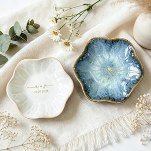 K&ouml;nnte beinhalten: Zwei blumenf&ouml;rmige Schmuckschalen aus Keramik. Eine ist gl&auml;nzend wei&szlig; mit goldenem Text a + g und 05.07.2026. Die andere hat eine reaktive blau-wei&szlig;e Glasur mit einem goldenen Herz und Text. Beide haben gewellte R&auml;nder und eine glatte Oberfl&auml;che.