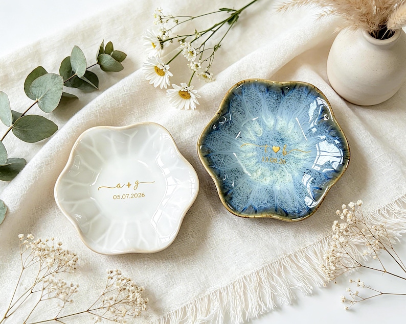 K&ouml;nnte beinhalten: Zwei blumenf&ouml;rmige Schmuckschalen aus Keramik. Eine ist gl&auml;nzend wei&szlig; mit goldenem Text a + g und 05.07.2026. Die andere hat eine reaktive blau-wei&szlig;e Glasur mit einem goldenen Herz und Text. Beide haben gewellte R&auml;nder und eine glatte Oberfl&auml;che.
