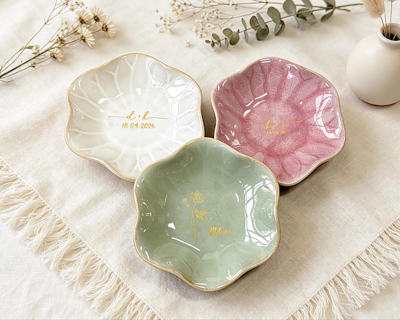 K&ouml;nnte beinhalten: Drei blumenf&ouml;rmige Schmuckschalen aus Keramik in Wei&szlig;, Rosa und Salbeigr&uuml;n mit einer rissigen Glasuroberfl&auml;che. Jede Schale ist mit goldfarbenen Schriftz&uuml;gen und Motiven verziert, darunter "d+k" mit "18.04.2026" sowie der Name "Melanie".