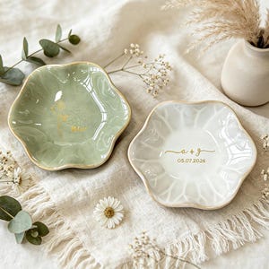 K&ouml;nnte beinhalten: Zwei gewellte Schmuckschalen aus Keramik mit goldener Beschriftung. Eine Schale ist salbeigr&uuml;n mit einem goldenen Blumenmotiv und dem Namen "Alice". Die andere Schale ist wei&szlig; mit dem goldenen Text "a + g" und dem Datum "05.07.2026".