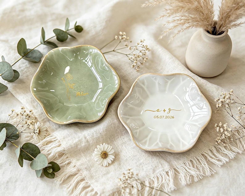K&ouml;nnte beinhalten: Zwei gewellte Schmuckschalen aus Keramik mit goldener Beschriftung. Eine Schale ist salbeigr&uuml;n mit einem goldenen Blumenmotiv und dem Namen "Alice". Die andere Schale ist wei&szlig; mit dem goldenen Text "a + g" und dem Datum "05.07.2026".