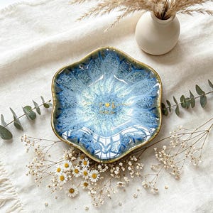 K&ouml;nnte beinhalten: Eine sechseckige Keramikschale mit einer reaktiven blau-wei&szlig;en Glasur und einem dunkelbraunen Rand. In der Mitte der Schale befinden sich goldene Schriftzeichen und ein kleines Herzsymbol. Die Schale hat einen gewellten Rand und eine glatte Oberfl&auml;che.