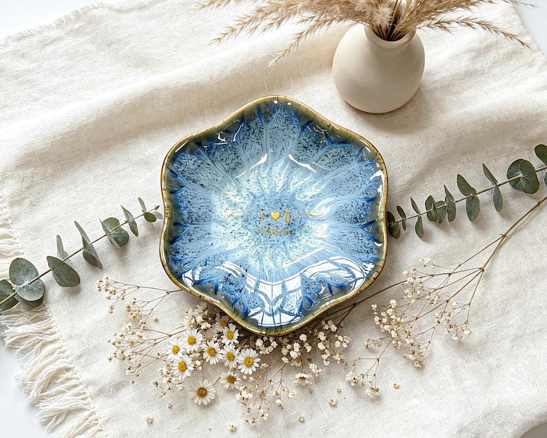 K&ouml;nnte beinhalten: Eine sechseckige Keramikschale mit einer reaktiven blau-wei&szlig;en Glasur und einem dunkelbraunen Rand. In der Mitte der Schale befinden sich goldene Schriftzeichen und ein kleines Herzsymbol. Die Schale hat einen gewellten Rand und eine glatte Oberfl&auml;che.