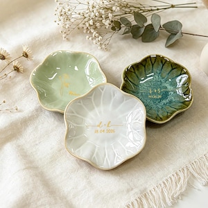 K&ouml;nnte beinhalten: Drei blumenf&ouml;rmige Schmuckschalen aus Keramik in Hellgr&uuml;n, Dunkelgr&uuml;n und Wei&szlig;. Jede Schale ist in der Mitte mit einem goldenen Monogramm und einem Datum versehen. Die wei&szlig;e Schale zeigt den Text d + h 18.04.2026, die dunkelgr&uuml;ne Schale s + s 07.06.26.
