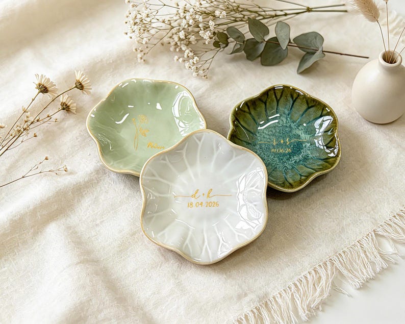 K&ouml;nnte beinhalten: Drei blumenf&ouml;rmige Schmuckschalen aus Keramik in Hellgr&uuml;n, Dunkelgr&uuml;n und Wei&szlig;. Jede Schale ist in der Mitte mit einem goldenen Monogramm und einem Datum versehen. Die wei&szlig;e Schale zeigt den Text d + h 18.04.2026, die dunkelgr&uuml;ne Schale s + s 07.06.26.