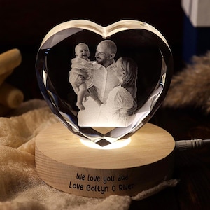 May include: A heart-shaped crystal photo featuring a family portrait, lit by a warm glow. The crystal is on a wooden base with the inscription "We love you dad Love Coltyn & River". A unique personalised gift.