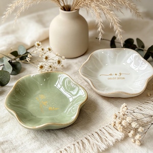 K&ouml;nnte beinhalten: Zwei kleine Keramikschalen mit gewelltem Rand. Eine Schale ist salbeigr&uuml;n mit einem goldenen Blumenmotiv, die andere ist wei&szlig; mit dem goldenen Text a + g und 05.07.2026. Beide Schalen haben eine glatte, gl&auml;nzende Oberfl&auml;che und goldene R&auml;nder.