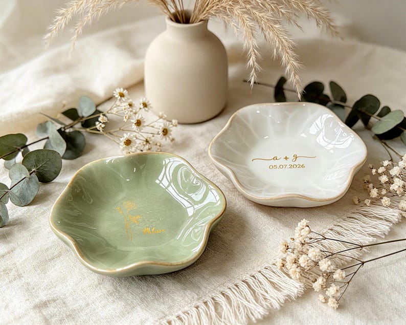 K&ouml;nnte beinhalten: Zwei kleine Keramikschalen mit gewelltem Rand. Eine Schale ist salbeigr&uuml;n mit einem goldenen Blumenmotiv, die andere ist wei&szlig; mit dem goldenen Text a + g und 05.07.2026. Beide Schalen haben eine glatte, gl&auml;nzende Oberfl&auml;che und goldene R&auml;nder.