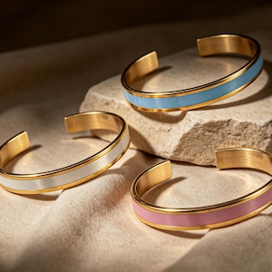 May include: Three open-ended cuff bracelets made of gold-toned metal. Each bracelet features a smooth, coloured inlay band running through the centre. The colours of the inlay bands are light blue, white, and soft pink, respectively.