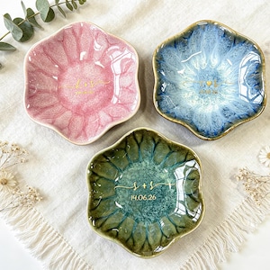 K&ouml;nnte beinhalten: Drei blumenf&ouml;rmige Schmuckschalen aus Keramik in Rosa, Blau und Gr&uuml;n mit gl&auml;nzender Reaktivglasur. Jede Schale ist in der Mitte mit goldfarbenen Buchstaben, Initialen und dem aufgedruckten Datum 14.06.26 versehen.