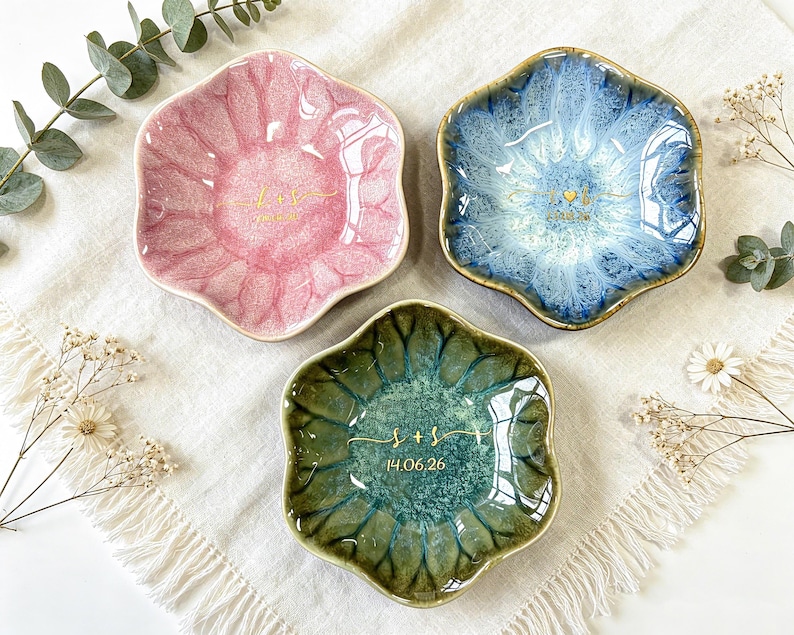 K&ouml;nnte beinhalten: Drei blumenf&ouml;rmige Schmuckschalen aus Keramik in Rosa, Blau und Gr&uuml;n mit gl&auml;nzender Reaktivglasur. Jede Schale ist in der Mitte mit goldfarbenen Buchstaben, Initialen und dem aufgedruckten Datum 14.06.26 versehen.