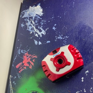 May include: A red and silver digital camera with the word "Digital" printed on the front. The camera is on a blue surface with white and red paint splatters.