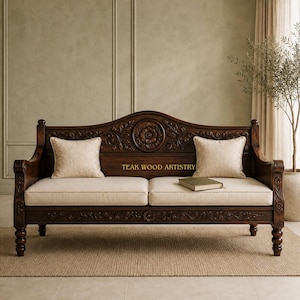 May include: A dark brown wooden sofa with intricate floral carvings on the backrest and frame. It features two beige upholstered seat cushions, two matching beige throw pillows, and a small book resting on the seat. The text TEAK WOOD ARTISTRY is visible.