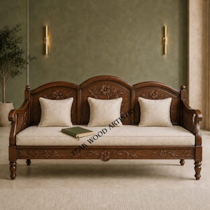 May include: A dark brown wooden sofa featuring intricate floral carvings on the backrest and frame. The sofa includes a long, light beige seat cushion and three matching square throw pillows. A small green book rests on the seat cushion.