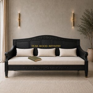 May include: A black wooden sofa with a curved backrest featuring carved floral details and the text TEAK WOOD ARTISTRY. The sofa includes a long off-white seat cushion and three matching rectangular throw pillows, with a small green book on the seat.