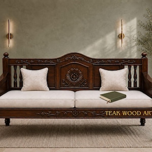 May include: A dark brown wooden sofa with intricate floral carvings on the backrest and frame. It features two off-white seat cushions, two matching square throw pillows, and a small green book resting on the seat. The text TEAK WOOD ARTISTRY is visible.