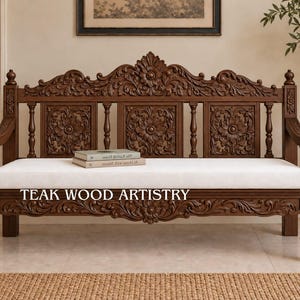May include: A dark brown wooden bench featuring intricate floral carvings on the backrest and frame, with a plain white rectangular seat cushion. The text TEAK WOOD ARTISTRY is printed in white on the front of the wooden frame below the cushion.