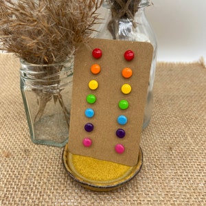 Peut inclure: Un ensemble de boucles d'oreilles clous arc-en-ciel sur une carte marron. Les boucles d'oreilles sont disposées en forme d'arc-en-ciel, avec du rouge, de l'orange, du jaune, du vert, du bleu, du violet et du rose.