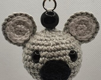 May include: A handmade crochet koala keychain. The koala has a gray body, black nose, and black eyes. The ears are gray with a lighter gray trim. A black bead is attached to the keychain chain.