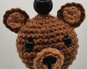 May include: A brown crochet teddy bear keyring with black bead eyes and nose. The bear's head is attached to a silver-coloured chain and a black bead. The keyring is a charming accessory.