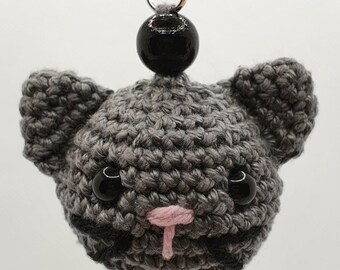 May include: A handmade crochet cat keychain in shades of gray. The cat's face features black button eyes, a pink nose, and a black stitched mouth. A black bead sits above the cat's head, attached to a silver keyring.