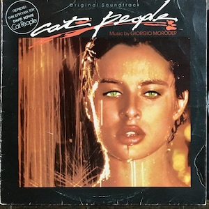 Puede incluir: Una portada de disco de vinilo negro con la cara de una mujer en el frente. La mujer tiene el pelo largo y oscuro y lleva una camisa blanca. El título del disco es "Cat People" y el texto "Original Soundtrack" está por encima del título. El texto "Music by GIORGIO MORODER" está por debajo del título.