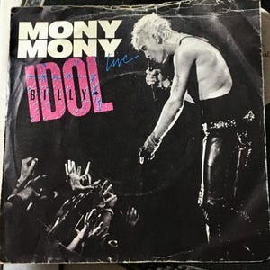 Puede incluir: Una foto en blanco y negro de Billy Idol actuando en vivo. La imagen muestra a Idol cantando en un micrófono con la mano levantada. El texto "Mony Mony" y "Billy Idol Live" es visible en la imagen.