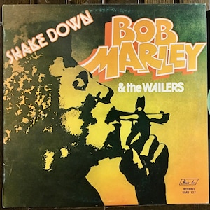 Könnte beinhalten: Ein Vintage-Vinyl-Plattencover von Bob Marley & The Wailers. Das Cover ist gelb, orange und grün mit einer schwarz-weißen Silhouette von Bob Marley, der eine Zigarette raucht. Der Albumtitel "Shake Down" steht in weißen Buchstaben oben auf dem Cover. Das Plattenlabel ist "Music-box" und die Katalognummer ist "SMB 127".