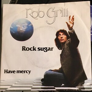 Può includere: Copertina di un disco in vinile vintage con Rob Grill. La copertina include una grafica del globo blu e bianco, il testo "Rock Sugar" e "Have Mercy". Un uomo è raffigurato che guarda in alto con la mano alzata.