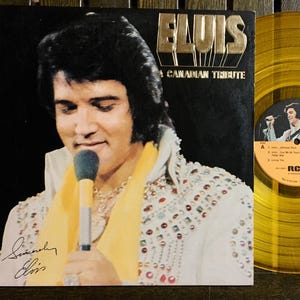 Peut inclure: Une pochette d'album vinyle noir et or représentant un portrait d'Elvis Presley chantant dans un microphone. Le titre de l'album est "Elvis: A Canadian Tribute".