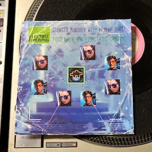 May include: A vinyl record cover featuring a blue and white design with a grid pattern. The cover has the text "Giorgio Moroder with Philip Oakey" and "Together in Electric Dreams" on it. There are multiple images of people on the cover.