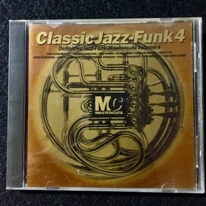 Puede incluir: Una caja de CD con fondo dorado y una imagen en negro y dorado de un trompa. El texto "Classic Jazz-Funk 4" está impreso en negro en la parte superior de la caja. El texto "Definitive Jazz-Funk Mastercuts volume 4" está impreso en negro debajo del título. El texto "MC Mastercuts" está impreso en negro en la parte inferior de la caja.