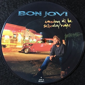 May include: A black vinyl record with a picture of a man sitting in front of a diner at night. The record is titled "Bon Jovi - Someday I'll Be Saturday Night".