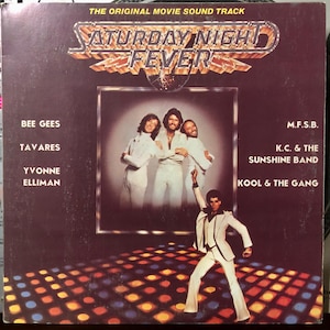 Può includere: Copertina dell'album della colonna sonora di Saturday Night Fever. La copertina presenta il titolo in oro, in un font stilizzato, sopra una foto dei Bee Gees e un uomo in abito bianco che balla su una pista da ballo. I nomi degli artisti sono elencati a sinistra e a destra.