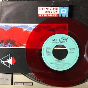 Pode incluir: Um disco de vinil vermelho de Depeche Mode, intitulado "Stripped", com uma sobreposição vermelha transparente a condizer. O disco está numa gira-discos, com a capa visível. A capa apresenta o nome da banda e uma paisagem.