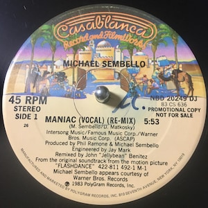 May include: A vintage vinyl record for Michael Sembello's song "Maniac (Vocal) (Re-Mix)". The record label features a desert scene with palm trees and a city. The text includes "Casablanca Record and FilmWorks" and "Promotional Copy Not For Sale".