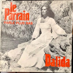 May include: Album cover featuring Dalida in a white dress, seated on rocks near the water. The text "Le Parrain" and "Parle Plus Bas" is in red, along with the artist's name, "Dalida."