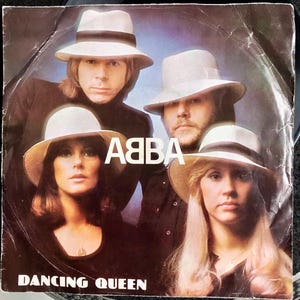 Op de afbeelding: Een vierkante vinylplatenhoes met een portret van vier personen die witte fedora-hoeden met donkere banden dragen. De groep is gekleed in donkere kleding. De tekst ABBA staat in het wit in het midden en DANCING QUEEN staat onderaan.