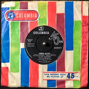 Pode incluir: Um disco de 45 RPM com um rótulo preto e uma capa listrada colorida. O disco é intitulado "Summer Holiday" por Cliff Richard e The Shadows. A capa tem o logotipo da Columbia e o texto "This record must be played at 45 RPM."